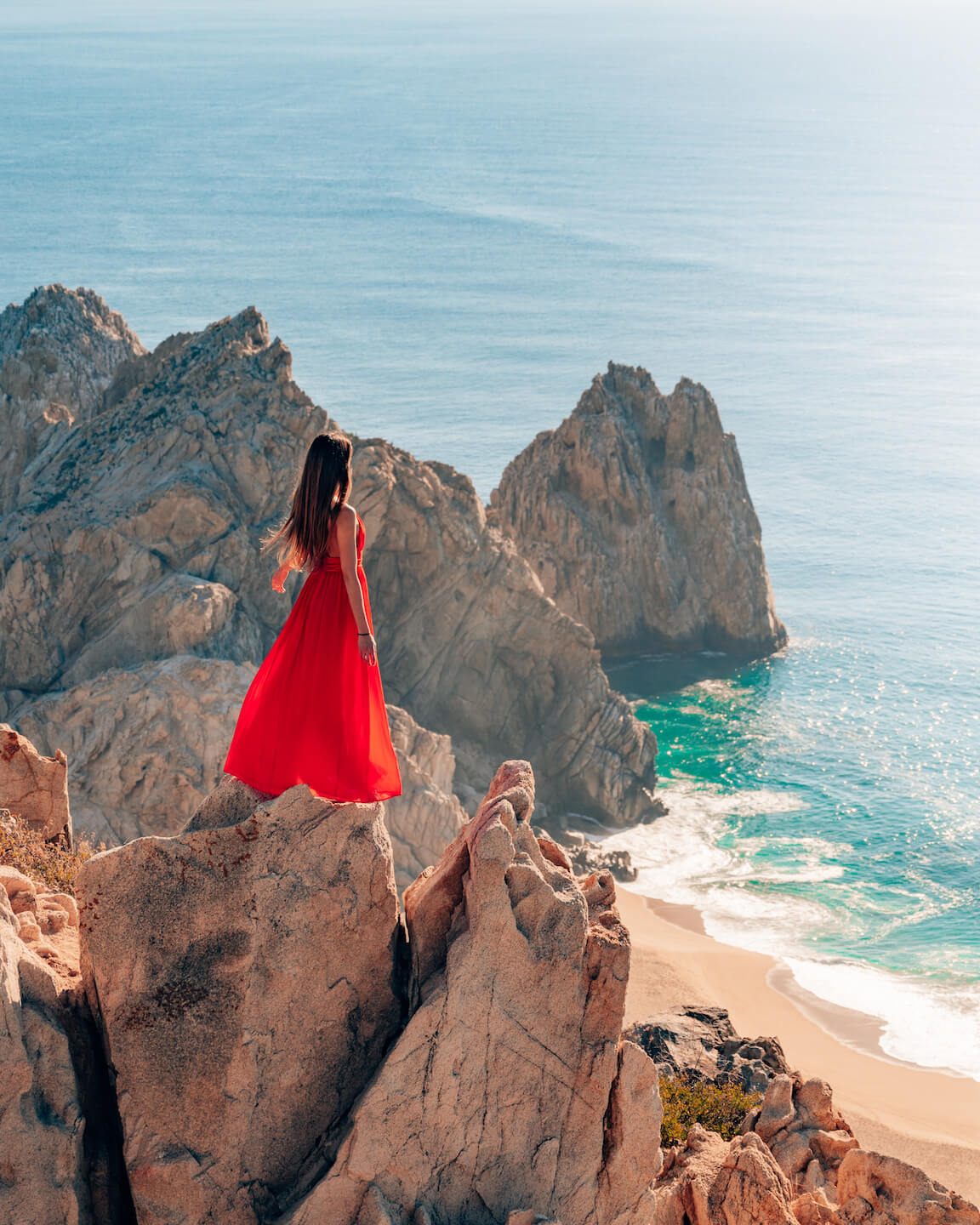 girl in red dress standing on rock and overlooking Beach in Cabo